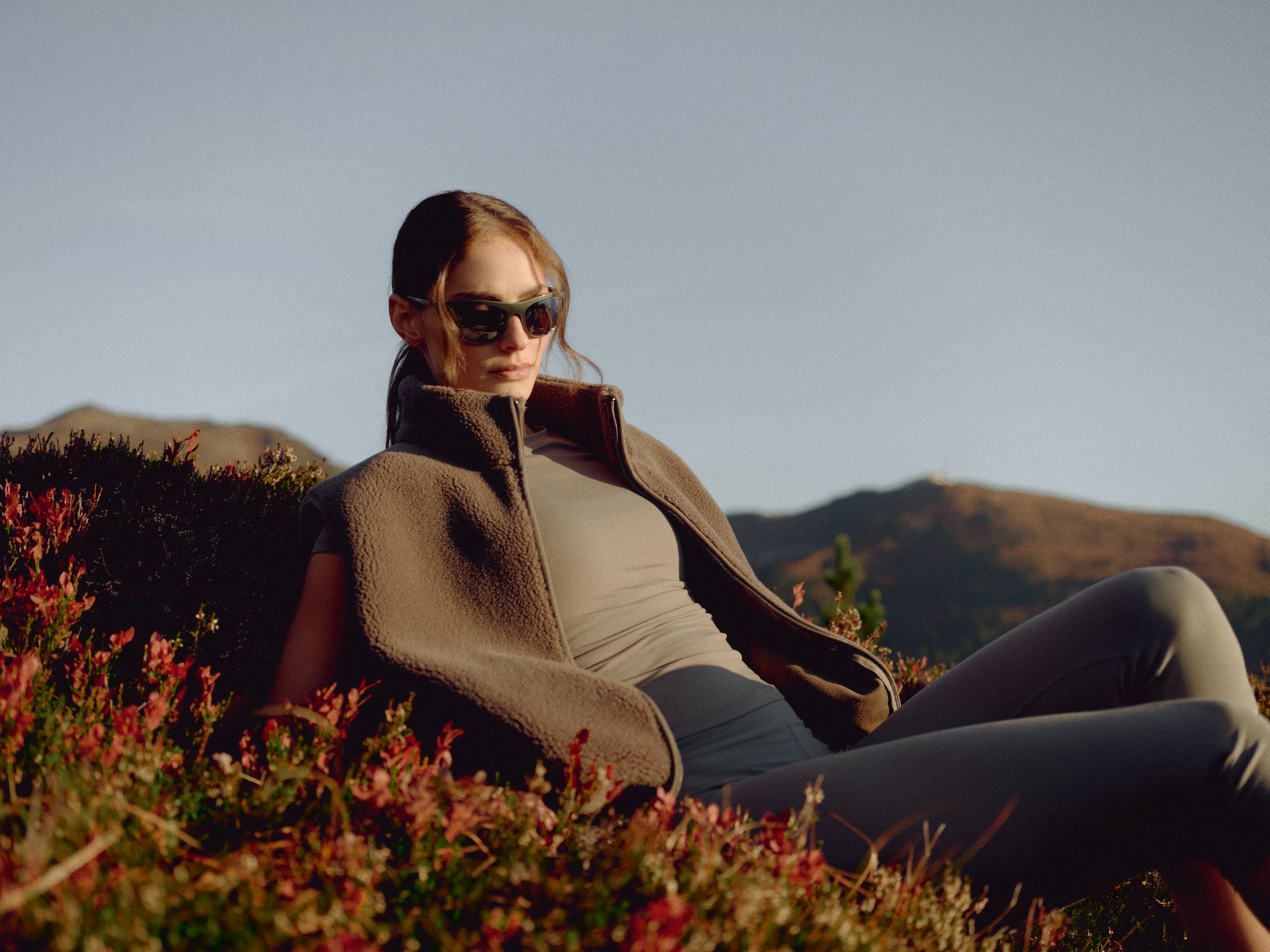 Woman wearing a khaki CASPARA Cloudfleece vest and sunglasses on a grassy hillside.